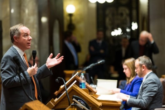 A man in a suit and tie speaks at a podium to a room of legislators